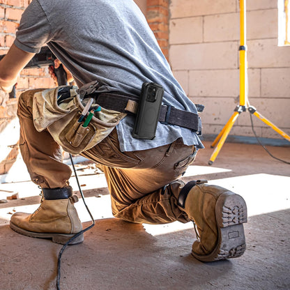 Construction worker kneeling at job site wearing Motivecase Bunker case for iPhone 15 Pro Max attached securely to tool belt clip holster.